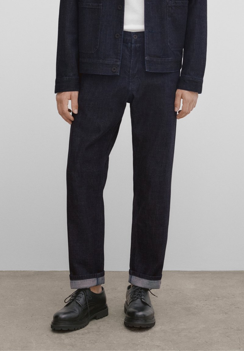 Tummansiniset farkkutakki ja housut, suoralla leikkauksella, napillinen sulkeminen ja käännetyt hihansuut. Yhdistetty mustiin nyörikenkiin.