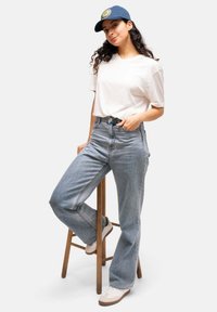Camiseta blanca de manga corta combinada con unos vaqueros de cintura alta de color azul claro y zapatillas beige. El modelo lleva una gorra azul con un logo redondo.