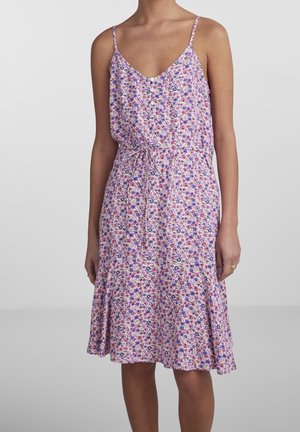 Kvinde iført ærmeløs, knælængde, blomsterprintet kjole med spaghetti stropper og bindebånd i taljen mod en ensfarvet baggrund.