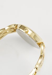 Gold-tone metal watch featuring a round case, smooth bezel, and a textured crown. The strap has multiple linked sections for flexibility.