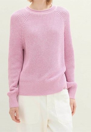 Kvinde iført en lyserosa strikket sweater med lange ærmer og hvide højttaljede bukser, stående mod en ensfarvet baggrund.