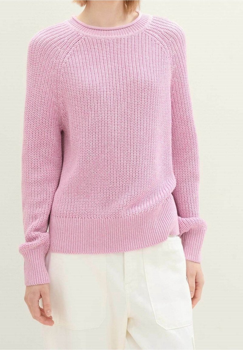 Femme portant un pull en maille rose clair à manches longues et un pantalon blanc taille haute, debout devant un fond uni.