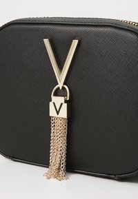 Borsa in pelle sintetica nera con un grande accento dorato a forma di "V" e un attacco con nappina a catena. Superficie testurizzata con chiusura a zip.