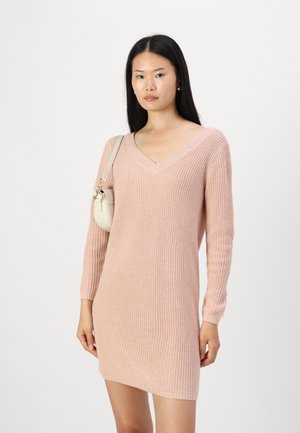 Robe en tricot rose avec un col en V et des manches longues. Présente un motif texturé et une silhouette ajustée, associée à un petit sac à main clair.