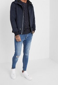 Giacca zip nera con cappuccio sopra un maglione blu a fantasia, abbinata a jeans azzurri chiari strappati e sneakers bianche.