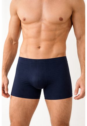 Torso maschile muscoloso che indossa boxer aderenti blu scuro tinta unita, in piedi con le braccia rilassate su uno sfondo bianco.