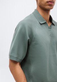 Άνδρας που φοράει κοντομάνικο μπλουζάκι πόλο σε απαλό πράσινο χρώμα με υφασμάτινες πλεκτές επιφάνειες και ριμπ γιακά και μανσέτες, σε απλό φόντο.