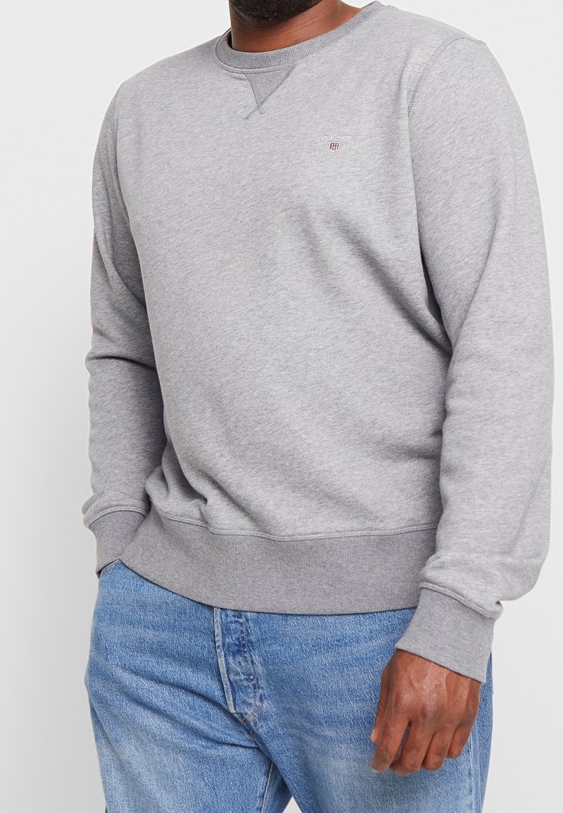Homme portant un sweat-shirt gris clair à manches longues avec un petit logo et un jean bleu, debout devant un fond uni.