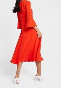 Robe midi rouge avec manches évasées et ourlet fluide, fabriquée en tissu lisse. Portée avec des chaussures blanches à talons hauts.