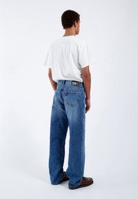 Blue denim jeans with a relaxed fit, featuring a faded finish, five pockets, and a black label on the waistband. White shirt is worn above.