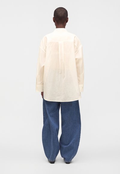 Personne debout de dos, portant une chemise ample crème à manches longues et un jean large bleu sur un fond clair uni.