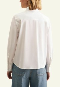Camicia bianca con bottoni, dalla vestibilità rilassata, con cucitura a yoke sulla schiena e maniche lunghe. Tessuto liscio con orlo arrotondato e polsini con bottoni.