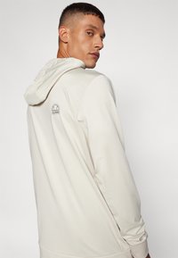 Krēmveida kapuci treneris ar logo uz muguras, gluda tekstūra, brīva piegriezuma un garām piedurknēm.