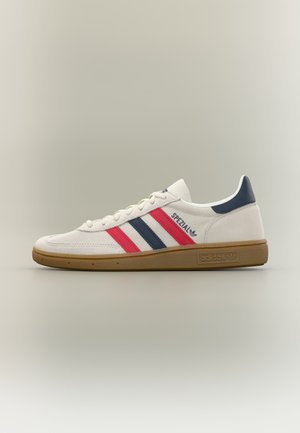 Άσπρο sneaker Adidas Spezial με κόκκινες και ναυτικές ρίγες, καφέ σόλα και ναυτικό ταμπάκι στη φτέρνα σε ουδέτερο φόντο.