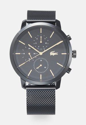 Montre en acier inoxydable noir avec un bracelet en maille, dotée d'un cadran sombre, de marqueurs d'heure en or, de sous-cadrans et d'un logo crocodile à la position 4 heures.