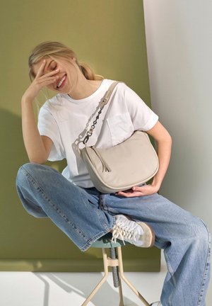 Junge Frau sitzt mit überkreuzten Beinen auf einem Hocker, trägt ein weißes T-Shirt, blaue Jeans, weiße Sneakers, hält eine beige Umhängetasche und lächelt, während sie ihre Hand an das Gesicht hält.