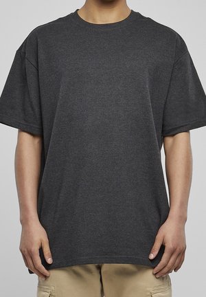 Osoba ubrana w prostą, ciemnoszarą, oversize koszulkę z krótkim rękawem i jasnobeżowe spodnie, stoi z ramionami rozluźnionymi wzdłuż ciała.