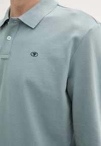 Lyseblå polo-shirt lavet af tekstureret stof, med krave og et lille logo på venstre side. To knapper på placket.