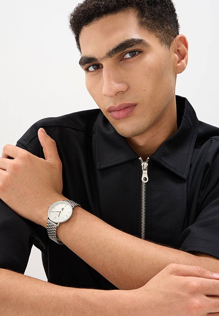 Orologio argento con quadrante bianco e bracciale a rete, indossato su un polso. Camicia nera con zip e colletto, modello con le braccia appoggiate sul petto.