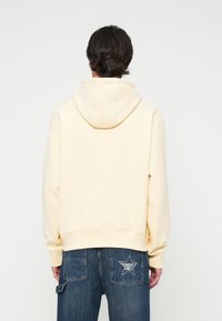 Sudadera con capucha en color amarillo pálido, con un tejido suave y texturizado, bolsillo tipo canguro y un corte relajado. Combinada con unos jeans de mezclilla azul que exhiben un diseño de estrellas.