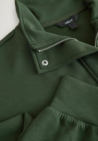 Dark green zip-up jacket with collar featuring a silver snap button and visible brand label "NEXT" inside the neckline.