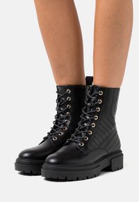 Botas de combate de cuero negro con ojales dorados, diseño de cordones y un detalle de raya vertical texturizada en el cañón. Suela gruesa y resistente.