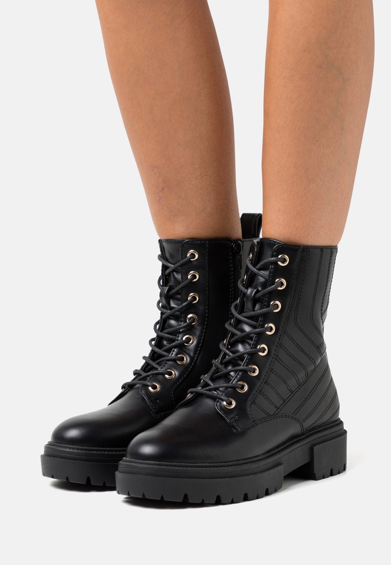 Botas de combate de cuero negro con ojales dorados, diseño de cordones y un detalle de raya vertical texturizada en el cañón. Suela gruesa y resistente.