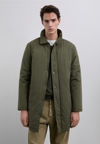 Olivengrøn quiltet jakke med en struktureret krave, lukkes med trykknapper og har sidelommer, kombineret med en grøn sweater og beige bukser.