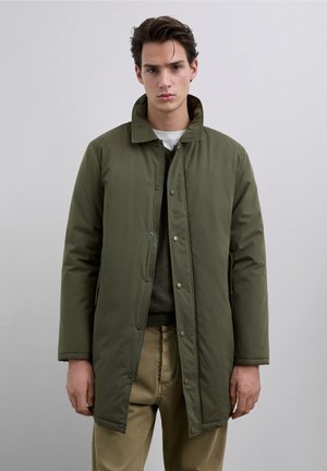 Chaqueta acolchada verde oliva con cuello estructurado, cierre de botones a presión y bolsillos laterales, combinada con un suéter verde y pantalones beige.