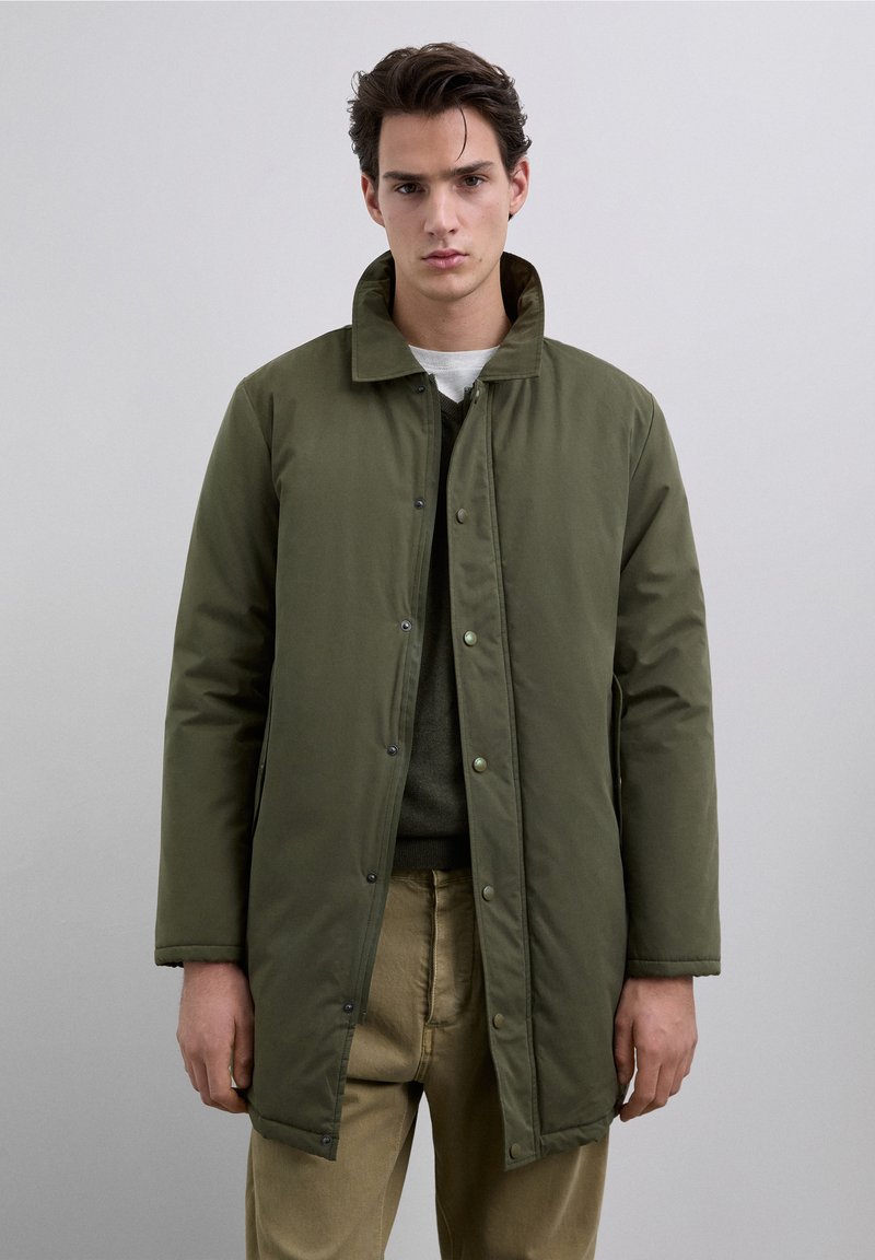 Olivengrøn quiltet jakke med en struktureret krave, lukkes med trykknapper og har sidelommer, kombineret med en grøn sweater og beige bukser.