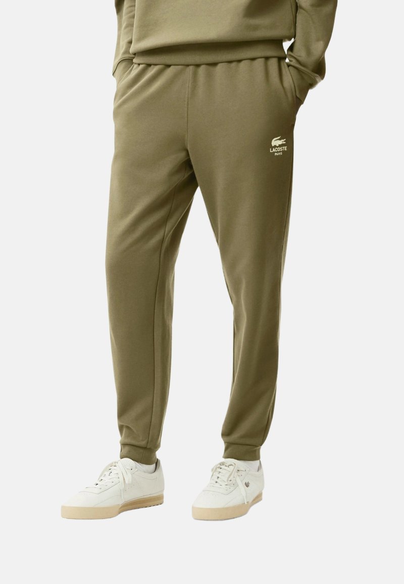 Pantalones deportivos verde oliva con cintura elástica y bolsillos laterales, con un logo blanco de Lacoste en el muslo izquierdo.
