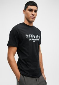 Młody mężczyzna w czarnej koszulce z białymi postaciami z kreskówek i napisem "MIŁOŚĆ DO NIEZNANEGO", stojący z rękami w kieszeniach.