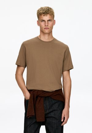 Blond man with short hair wearing a brown short-sleeve T-shirt, blue jeans, and a tied dark brown sweater around his waist.