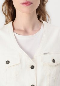 Hvit denimjakke med to brystlommer, sølvfargede knapper og V-hals design. Bært over en enkel hvit t-skjorte.