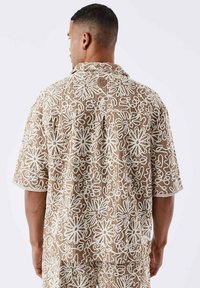 Camisa de manga corta con un fondo marrón claro y patrones florales y líneas abstractas en blanco, diseño con cuello y un pequeño parche con la marca en la parte posterior.