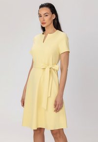 Robe jaune avec un corsage ajusté, des manches courtes, un décolleté en V et une ceinture à nouer à la taille. Tissu lisse, jupe arrivant au genou.
