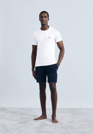 Camiseta blanca de manga corta con un pequeño logo negro, combinada con pantalones cortos azul marino, ambos hechos de tela suave. De pie sobre una superficie gris suave.
