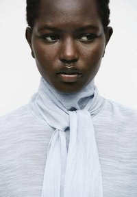 Blusa azzurro chiaro, con texture e colletto a sciarpa annodato, primo piano del viso che mostra una pelle liscia e un'espressione neutra.