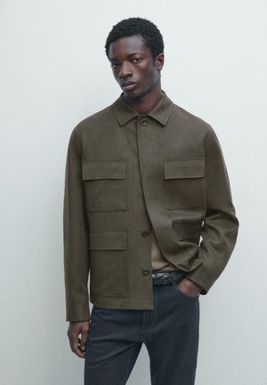 Uomo con giacca verde oliva a bottoni con quattro tasche con patta, maglione beige, pantaloni blu scuro, in piedi con una mano in tasca contro uno sfondo semplice.