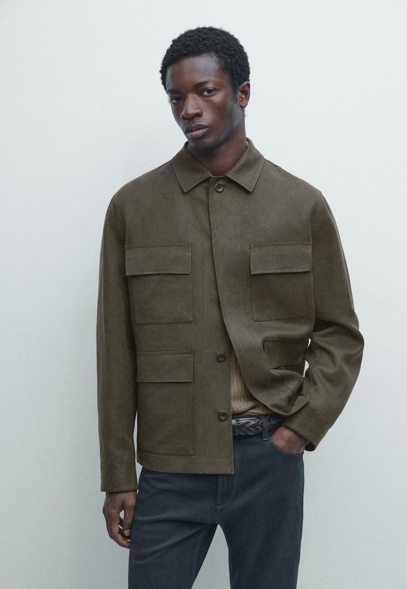 Mann trägt eine olivgrüne Jacke mit Knöpfen und vier Klappentaschen, einen beigen Pullover und dunkelblaue Hose, steht mit einer Hand in der Tasche vor einem einfarbigen Hintergrund.