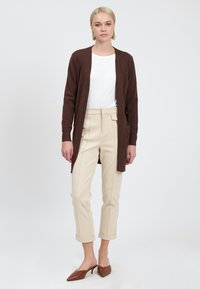 Donkerbruine cardigan over een witte top, gecombineerd met beige high-waisted broeken met opgerolde mouwen. Donkere puntige mules maken de outfit compleet.
