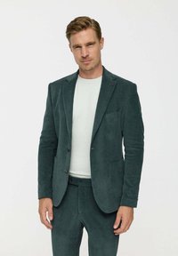 Sötétzöld kordbársony öltöny, ribbed textúrával, jól szabott blézerrel, két gombbal, és hozzáillő nadrággal. Alatta egyszerű, kerek nyakú ing.