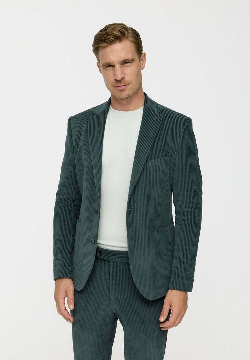 Sötétzöld kordbársony öltöny, ribbed textúrával, jól szabott blézerrel, két gombbal, és hozzáillő nadrággal. Alatta egyszerű, kerek nyakú ing.