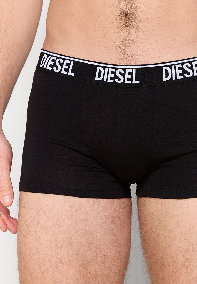 Männlicher Oberkörper und Oberschenkel tragen schwarze Diesel-Boxershorts mit einem logo-markierten Bund vor einem schlichten hellen Hintergrund.