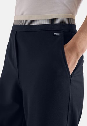 Nahaufnahme einer Person, die eine schwarze Hose mit einem beigefarbenen und schwarzen gestreiften elastischen Bund trägt und deren Hand in der rechten Tasche ruht.