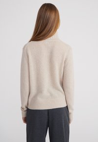 Beige Pullover mit hohem, geripptem Kragen und geripptem Saum. Weiche, texturierte Strickware. Kombiniert mit dunkelgrauen, weit geschnittenen Hosen.
