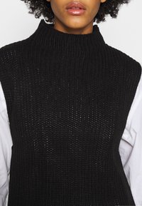 Schwarzes, gestricktes, ärmelloses Oberteil mit hohem Kragen und strukturiertem Muster. Wird über einem weißen Hemd getragen und hat eine lockere, entspannte Passform.