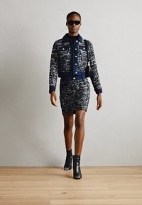Veste en denim texturée bleu et blanc et jupe près du corps, associées à des bottines noires et des lunettes de soleil, présentées dans un cadre minimaliste.