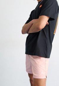 Person som står med armarna i kors, iklädd en svart t-shirt och rosa träningsbyxor med en vit logotyp på vänster ben mot en neutral bakgrund.