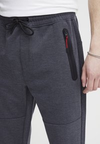 Tmavě šedé tepláky s texturovaným herringbone vzorem, s černou kapsou na zip a stahovacími šňůrkami v pase.